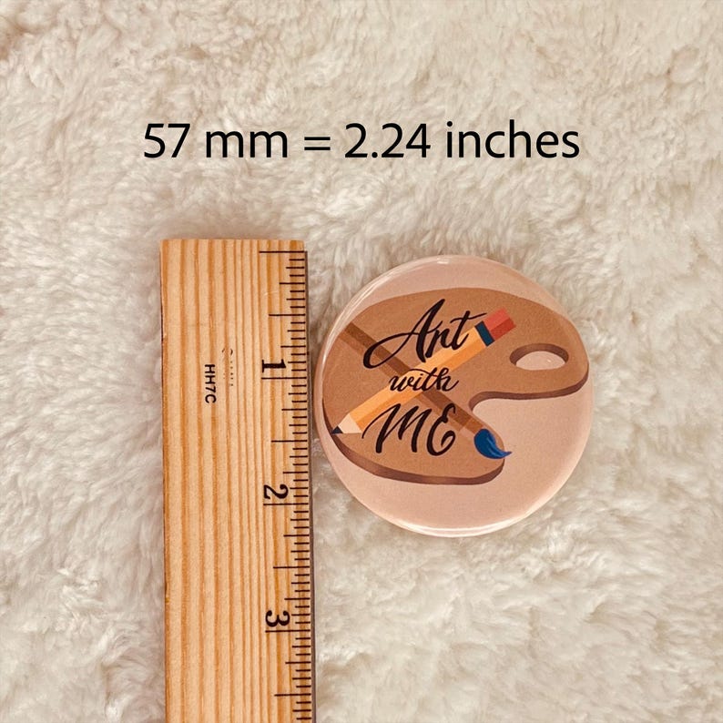 May include: A 2.24 inch round button with a pink background and a brown paint palette with a pencil and paintbrush. The text "Art with Me" is written in black cursive.