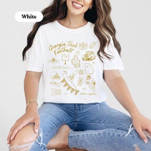 May include: White t-shirt with a gold graphic design featuring the text "Georgia Tech Football" and various illustrations. The design includes a football, stadium, and other related images. The shirt is a classic crew neck style.