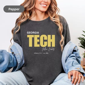 May include: Pepper-colored sweatshirt featuring the text "GEORGIA TECH" in bold yellow letters. Below the text, it reads "Yellow Jackets" in a script font, with "Atlanta, GA | est. 1885" underneath. The sweatshirt is a crew neck style.