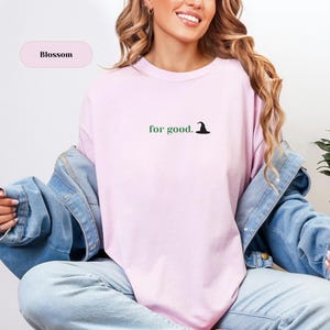 May include: A pale pink sweatshirt featuring the words "for good." in green, accompanied by a small black witch hat graphic. The word "Blossom" is in a pink oval. The sweatshirt is worn with a denim jacket.