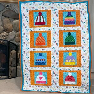 May include: A colorful quilt with a white background and a pattern of small blue and white hats. The quilt is divided into nine squares, each with a different colored hat with various designs. The hats are orange, green, purple, blue, and pink.