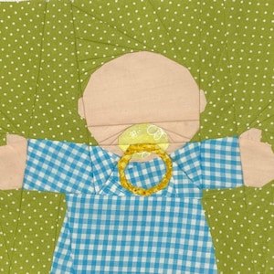 Op de afbeelding: Een blauwe en witte gingham babypop met een roze gezicht en een gele speen. De pop staat op een groene achtergrond met witte stippen.
