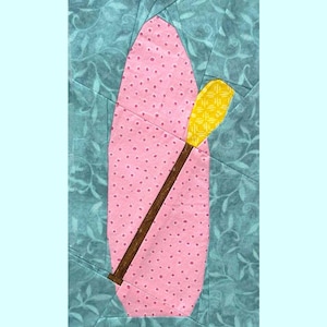 Könnte beinhalten: Ein Stoffdesign mit Boot und Paddel, mit einem rosa Boot mit kleinen Punkten und einem gelben Paddel. Das Boot und das Paddel sind vor einem türkisfarbenen Hintergrund mit floralem Muster angeordnet. Dies ist ein Quiltblock.