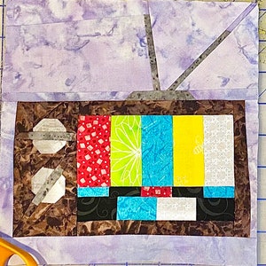 Puede incluir: Una pieza de arte textil hecha a mano que representa un televisor antiguo. La pantalla muestra barras de colores, con un fondo morado veteado y un marco marrón. La antena es gris.