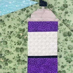 Puede incluir: Un bloque de patchwork de tela con un diseño de faro. El faro tiene un cuerpo morado y blanco, una parte superior gris y un acento negro. El fondo es un estampado floral verde, con una sección azul claro que representa el cielo.