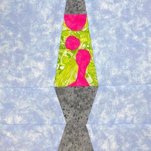 Puede incluir: Una lámpara de lava verde, amarilla y rosa con una base gris. La lámpara está hecha de tela y tiene un diseño acolchado.