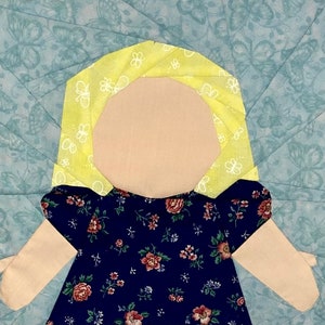 May include: A fabric doll with a light tan face and arms, wearing a navy blue dress with a floral pattern. The doll has yellow hair and is set against a light blue background with a butterfly pattern. The doll is a handmade craft.