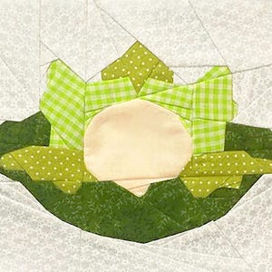 Puede incluir: Un bloque de edredón hecho a mano con un diseño estilizado en verde y blanco. El bloque presenta una forma central de color crema rodeada de hojas verdes y acentos en lunares y cuadros de vichy. El fondo es un estampado floral gris claro.