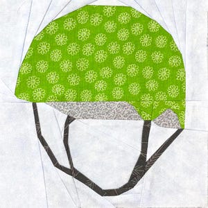Pode incluir: Um bloco de colcha de tecido com um capacete de equitação estilizado. A parte superior do capacete é verde brilhante com um padrão floral branco. A aba é cinza com um padrão floral e as correias são cinza escuro. O fundo é branco.