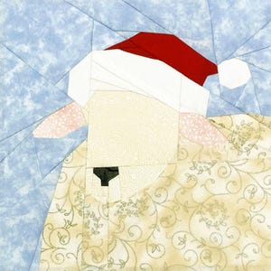 Op de afbeelding: Een stoffen kunstwerk dat een schaap met een kerstmuts afbeeldt. Het schaap heeft een crèmekleurig gezicht, een zwarte neus en een beige jas met een patroon. De kerstmuts is rood en wit, tegen een lichtblauwe achtergrond.