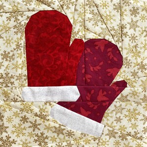 以下が含まれることがあります： 白いトリムが付いた2つの赤いミトンが特徴の、キルトのクリスマスをテーマにしたブロック。ミトンは、雪の結晶模様の金色の生地を背景に配置されています。ミトンには模様があります。