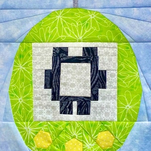 Pode incluir: Um bloco de colcha colorido com um design central de uma forma pixelizada em azul marinho e branco, sobre um fundo cinza e branco padronizado. A forma está contida dentro de um círculo verde com um padrão floral branco e três formas hexagonais amarelas na parte inferior.