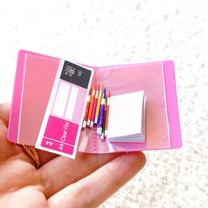 May include: A miniature pink A4 Clear File folder, open to reveal a small notepad and a collection of colorful pencils. The folder also contains a white and black label with the number 9. The overall theme is stationery.