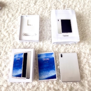 May include: A new tablet with a white frame and a blue screen displaying the time 00:00. The tablet is shown with its packaging, a charger, and a white paper bag labeled "Tablet."