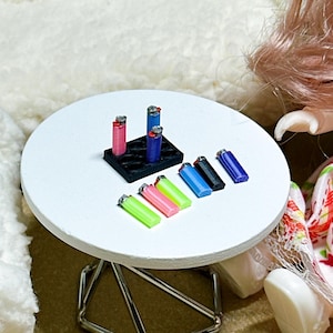 May include: A collection of miniature plastic lighters in various colors including neon green, pink, blue, and black. Some lighters are standing upright in a small black rectangular holder with circular slots, while others are lying flat.