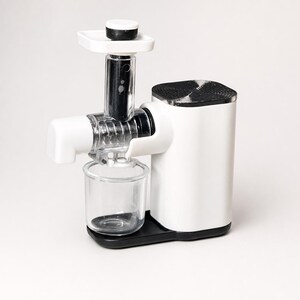 May include: A white and black electric juicer featuring a vertical feed chute with a white plunger, a clear plastic auger housing, and a small clear collection cup positioned on a black base. The main motor housing is white with a black top cover.