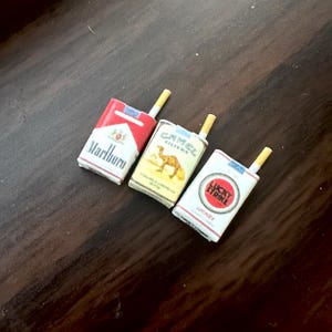 May include: Miniature cigarette packs of Marlboro, Camel, and Lucky Strike brands. Each pack is white with red accents and features a lit cigarette. The Camel pack displays a camel graphic, while the Lucky Strike pack has a red circle.