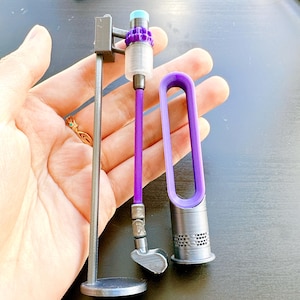May include: Miniature Dyson vacuum cleaner toy in purple, gray, and silver. The toy includes a stand, a handle, and a fan-shaped component. The vacuum has a cylindrical body with a purple top and a silver base. The toy is held in a hand.