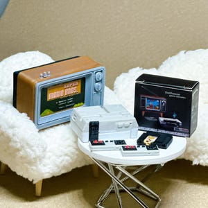 May include: A miniature brown and gray television set displaying Super Mario Bros, a gray game console, two small controllers, a black game cartridge, and a black product box labeled Dollhouse game consoles on a white circular table.