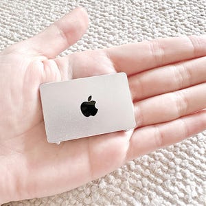 May include: A silver-colored laptop with a black Apple logo. The laptop is small and compact, and it is being held in a person's hand.