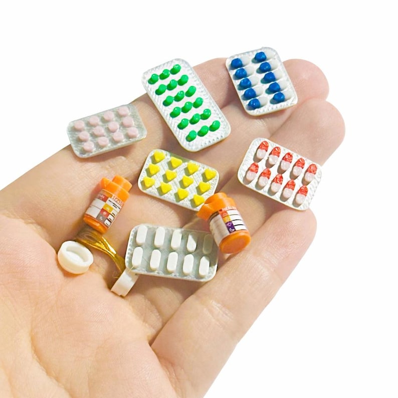 May include: Miniature medicine blister packs and pill bottles in various colors. The packs contain green, blue, pink, yellow, and red pills. The orange pill bottles have white labels. The items are small and likely for a dollhouse or craft project.