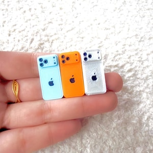 May include: Three miniature cell phones in light blue, orange, and white. Each phone has a black Apple logo and a camera array. The phones are held in a hand.