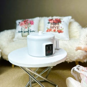 May include: A miniature white electric rice cooker with a digital control panel displaying the number 90. The appliance features a cylindrical body, a lid with a small knob, and a side handle, positioned on a small white round folding table.