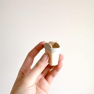 May include: A miniature, off-white takeout container with a hinged lid, held in a hand against a plain background. The container has a light beige body and a white rim and lid.