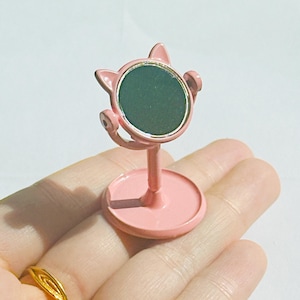 May include: A pink cat-shaped phone stand with a circular green screen. The stand has a round base and a stem that supports the screen. The cat ears and the stand are pink.