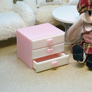 May include: A miniature pink storage unit with three white drawers, each featuring a small pink heart-shaped knob. The bottom drawer is pulled open, revealing the interior. The unit has a smooth, matte finish and a rectangular, boxy structure.