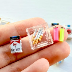 May include: Miniature Marlboro cigarette pack, cigarettes, lighters, and other accessories. The Marlboro pack is red and white. Two lighters are pink and neon green. The cigarettes are white with yellow filters.