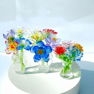 May include: A collection of colorful glass flower bouquets in small clear vases. The flowers include blue, yellow, red, and purple blooms. The vases are on a white surface.