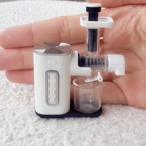 May include: A miniature white and silver juicer with a clear plastic container and a black base. The juicer has a vertical design with a top feeding chute and a side spout. The device has four buttons on the front.
