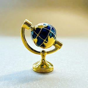 May include: A miniature globe ornament featuring a shiny gold-toned metal stand and a rotating sphere. The globe displays deep blue oceans with raised, polished gold-toned landmasses and a grid of thin gold lines representing latitude and longitude.
