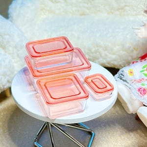 May include: A set of four clear rectangular food storage containers with orange lids. The containers vary in size, featuring transparent bodies and matching orange snap-on lids. These miniature containers are arranged in a cluster on a surface.