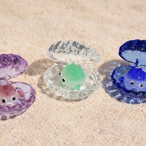 May include: Three glass seashell ornaments, each with a small sea creature inside. The shells are in purple, clear, and blue. The sea creatures are pink, green, and blue. The ornaments are on a sandy surface.