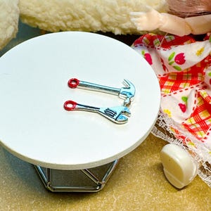 May include: Two miniature metal tool charms, a hammer and an adjustable wrench, both featuring silver-toned bodies and distinct red-painted circular ends on their handles, resting on a small, round, white tabletop surface.