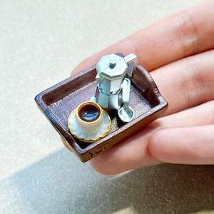 May include: A miniature brown rectangular tray holding a silver-toned moka pot, a small white coffee cup with a gold rim filled with dark liquid, and a matching silver-toned spoon. The set is crafted in a small scale for display or collection.