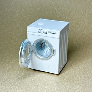 May include: A miniature white washing machine with a silver-rimmed circular door that is open. The front panel features a small control knob and printed text settings. The interior drum is visible, showing a perforated pattern in a light gray finish.