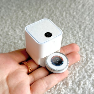 May include: A white, square-shaped device with a textured top and a small black circle. A smaller, round device with a screen displaying numbers and a silver rim is next to it. The devices are held in a hand.