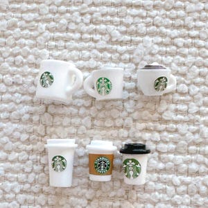 May include: Six miniature white ceramic Starbucks mugs with the Starbucks logo and a green mermaid. There are also two miniature Starbucks to-go cups, one with a brown paper sleeve and one with a black lid.