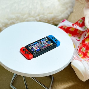 May include: A miniature handheld video game console featuring a black central screen displaying a grid of colorful game icons, flanked by a detachable red controller on the left and a detachable blue controller on the right side of the device.