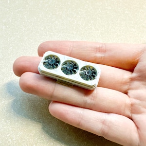 May include: A small, rectangular white component featuring three circular black fan-like structures arranged in a row. The object has a layered, tiered edge design and a small gold-colored connector tab visible on the bottom side of the frame.