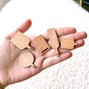 May include: A collection of small, wooden cutting boards in various shapes. The boards are light brown with a natural wood grain. Some have handles, and one is round. The boards are arranged in a hand.