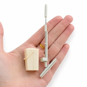 May include: Miniature cleaning set featuring a cream-colored bucket with a handle, a mop with a silver pole, and a separate squeegee. The set is designed for dollhouses or miniature displays. The items are held in a hand.