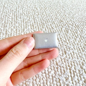 May include: A miniature silver laptop with the Apple logo on the lid. The small computer is held in a hand against a textured, off-white background. The laptop is a toy or decorative item.