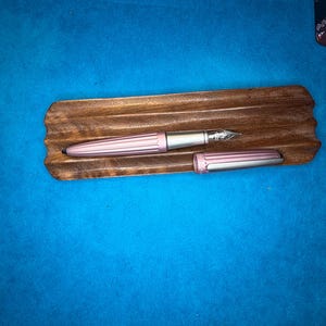 Puede incluir: Una pluma estilogr&aacute;fica rosa con plum&iacute;n y detalles plateados, colocada sobre un soporte de madera para bol&iacute;grafos. El capuch&oacute;n de la pluma est&aacute; quitado, revelando el plum&iacute;n. El soporte es de color marr&oacute;n oscuro y el fondo es azul liso.