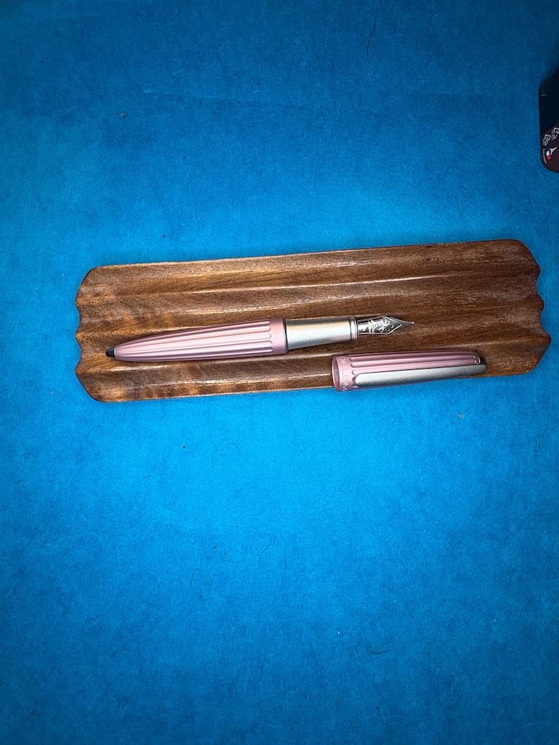 Puede incluir: Una pluma estilogr&aacute;fica rosa con plum&iacute;n y detalles plateados, colocada sobre un soporte de madera para bol&iacute;grafos. El capuch&oacute;n de la pluma est&aacute; quitado, revelando el plum&iacute;n. El soporte es de color marr&oacute;n oscuro y el fondo es azul liso.