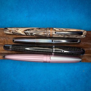 Puede incluir: Cuatro plumas estilogr&aacute;ficas de varios dise&ntilde;os y colores se muestran sobre una superficie de madera sobre un fondo azul. Las plumas incluyen una pluma veteada crema y marr&oacute;n, una pluma plateada, una pluma de fibra de carbono negra y una pluma rosa.