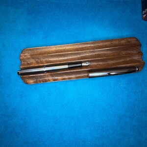 Puede incluir: Dos plumas estilogr&aacute;ficas plateadas descansan sobre un portal&aacute;pices de madera. Las plumas tienen un dise&ntilde;o elegante y moderno con detalles en negro. El soporte es de madera oscura con forma natural, sobre fondo azul.